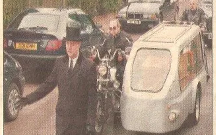 Motorcycle hearse carrying coffin down the road. Motor bike being led by funeral director wearing a frock coat and top hat.